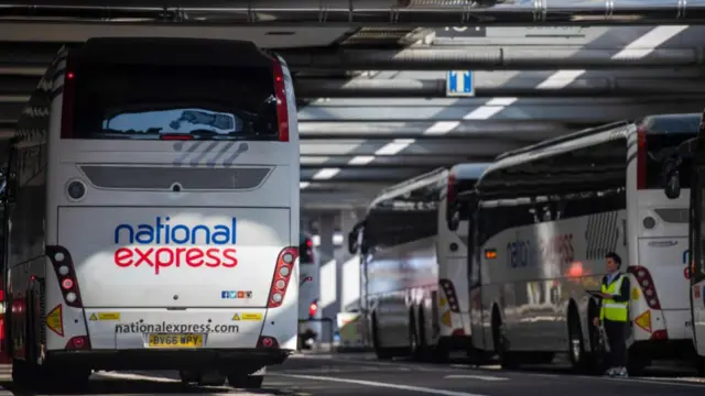 National Express coaches at bus station ready for departure. A coach is in the foreground on the left side of the picture with two more parked on the right, a third slightly visible to the far right of the frame. A man in a hi-vi jacket stands next to a coach