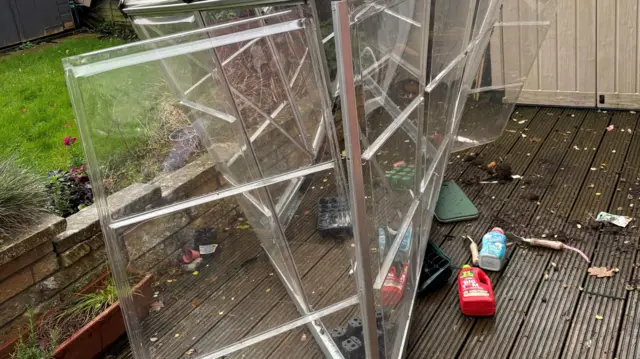 A crumpled greenhouse stands on a patio, clearly damaged