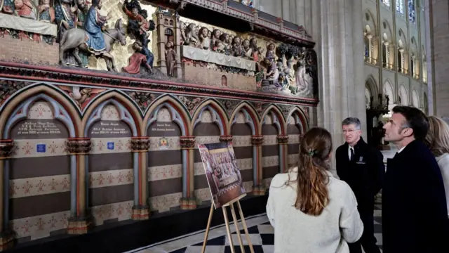 Macron stands in front of a sculptured wall. The bottom half of the wall has arches and the top half has people calved depicting scenes from the life of Jesus Christ. The wall is painted in earthy tones, including brown, dark red, pale blue, light yellow, and lilac