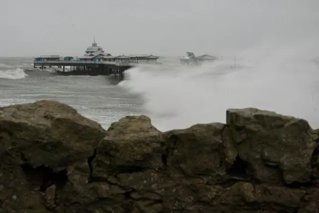 Wider shot of Llandudno pier in North Wales, with rock wall in foreground. One of the huts has been blown over into its side, resting on another hut. Large wave takes up about half of the image. Stormy waves and grey skies.