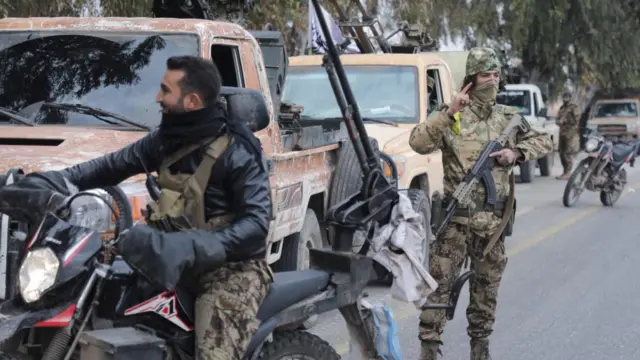 Members of Syrian rebel groups, one on a motorbike on left, a man in uniform holding a gun in the middle with vehicles covered in dust in the background