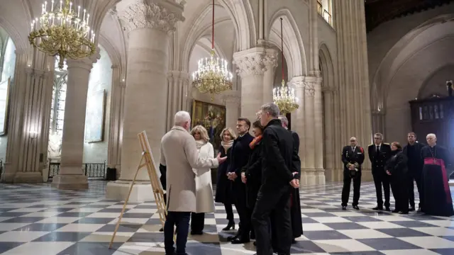 Macron stands with other people as a man talks to them. They are stood on a white and black checkered floor with golden chandeliers and cream-colours arches in the background