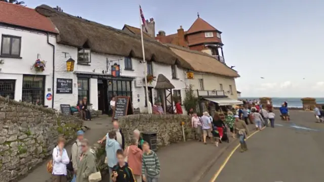 A thatched hotel with other thatched builidngs nearby and crowds of people with sea behind.