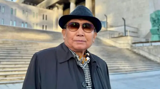 Koh Jae-hak. Elderly man wearing a dark grey hat, sunglasses, black windbreaker and chequered shirt in front of a set of stone steps