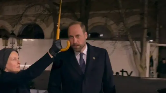 Prince Wiliam arrives at Notre Dame under the rain. A woman holds up an umbrella to cover him