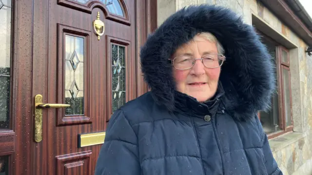 A woman standing outside her front door in a large coat with the hood up