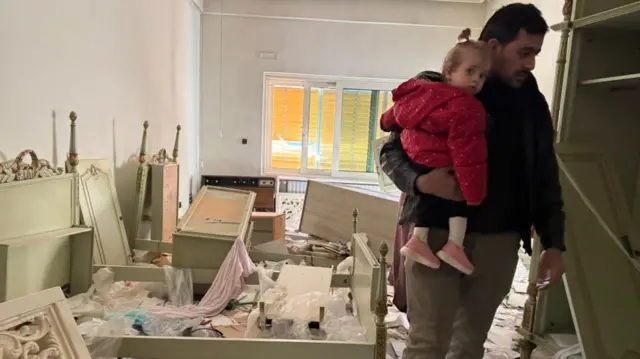 A man stands holding a little girl in a room in disarray