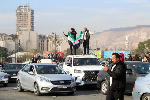 People celebrate in Damascus. Men holding the syrian rebel flag stand on top of a car with their fists in the air.