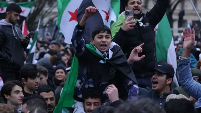 A boy sits on a man's shoulders, draped in the Syrian opposition flag and raising his fist