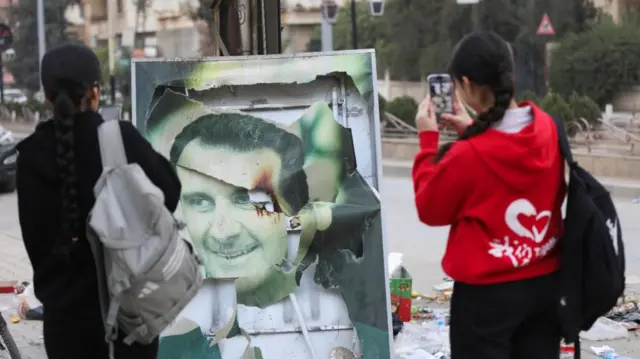 Women use their mobile phones near a damaged picture of Syrian President Bashar al-Assa