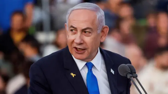 Benjamin Netanyahu in a suit and blue tie speaking into a microphone with blurred crowed in the background