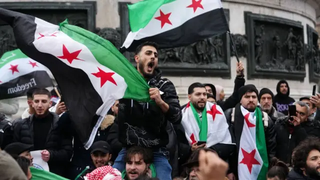 A man sits on another's shoulders amid a crowd, waving the Syrian opposition flag