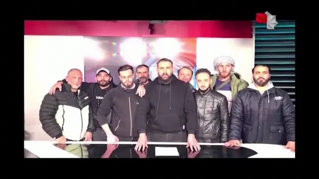 Syrian anti-government rebels stand in a television studio. There are nine people, all male, wearing hooded jumpers and jackets.