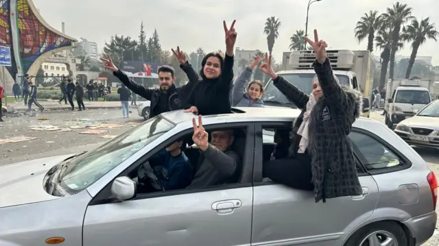 A number of people lean out of a car making peace signs with their hands