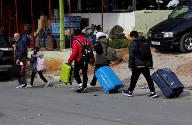 Four adults and one child, all wearing winter coats, carrying backpacks and suitcases walk along a street with parked car in background