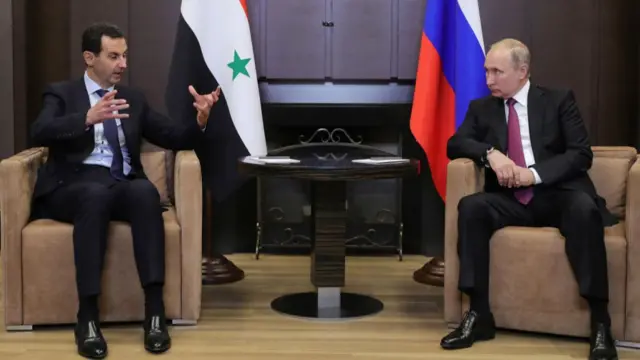 Bashar al-Assad (L) sitting down on beige armchair as he gestures with his hands while speaking to Vladimir Putin (R) siting on identical chair. Both are wearing dark suits. Next to Assad is a Syrian flag (red top, white centre with two green stars and black bottom), next to Putin a Russian one (white top, blue centre, red bottom)