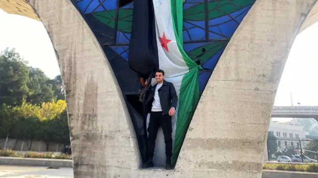 A man holds a gun as he stands in front of a Syrian opposition flag