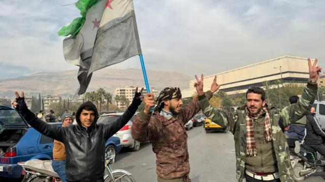 Three men hold their hands in the air, making a 'V' for victory sign, with the rebel flag held high