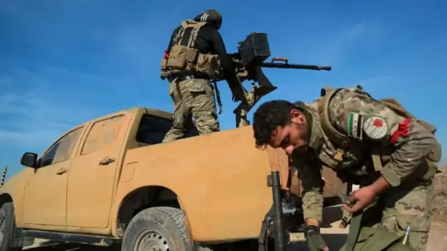 An HTS soldier stands on the back of a truck with a large gun mounted on the back, while another stands nearby with a weapon