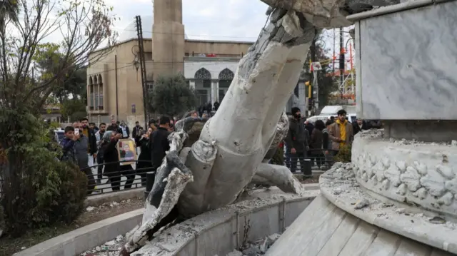 People stand near a damaged statue of former Syrian president Hafez al-Assad