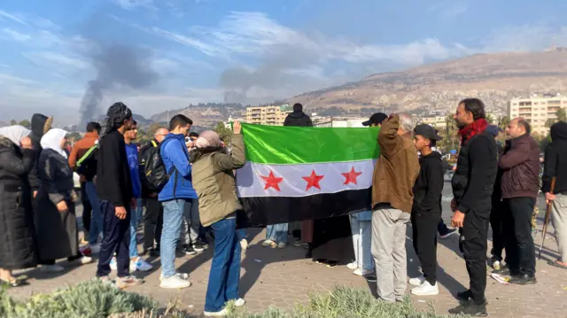 A group of people gathered facing away from the camera - a few hold up a Syrian opposition flag