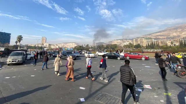 People file across a road as smoke rises in the distance