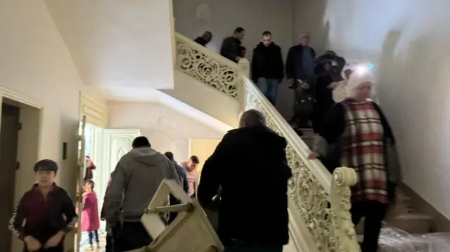 People file down the stairs, one takes a photo, another holds a piece of furniture