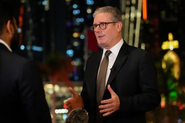 UK PM Keir Starmer in a dark suit, hands gesturing, as he talks to a reporter, partially seen from behind holding a microphone, with a blurred night time scene of lit up buildings in the background