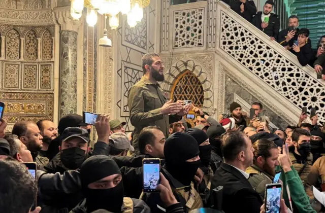 Group of people, some in black face masks, surround HTS leader Abu Mohammed al-Jawlani who stands slightly elevated, as he speaks, hands gesturing. People record scenes on their phones, and a group of people hang over the side of a set of stairs in the background