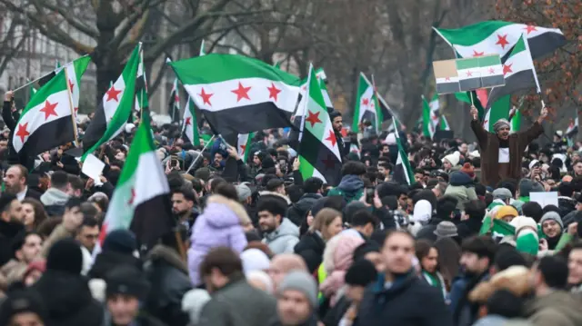 Syrians in Berlin celebrate the fall of the Assad regime. Large crowds of men and women wave the Syrian opposition flag (green top, white centre with three red stars and black bottom)