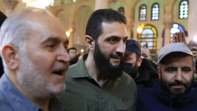 Abu Mohammad al-Jawlani walking in green fatigues surrounded by people during a visit to Great Umayyad Mosque in Damascus