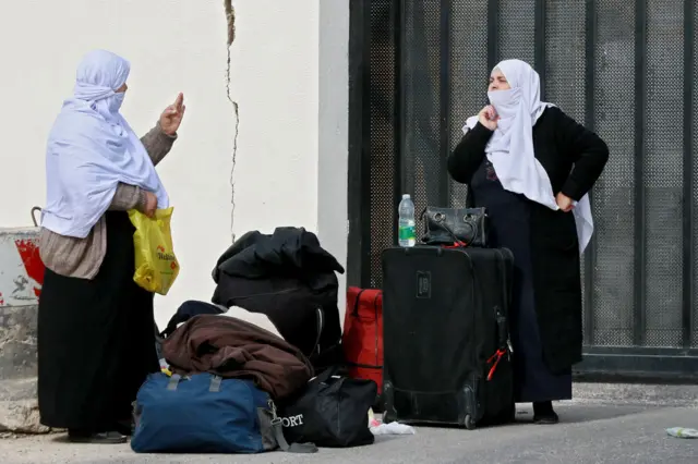 Two women, wearing white headscarves, stand here suitcases and bags with a wall and large gate behind them