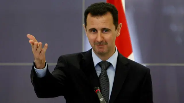 Bashar al-Assad gesture with one hand raised, palm up, while speaking in front of a microphone