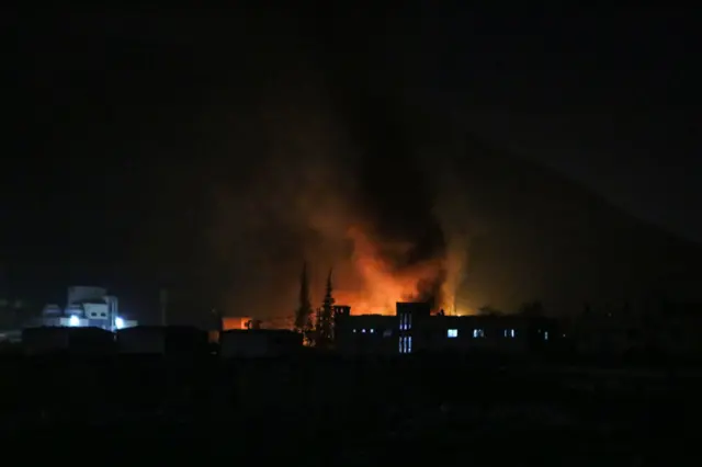 Smoke and fire is seen behind buildings and trees