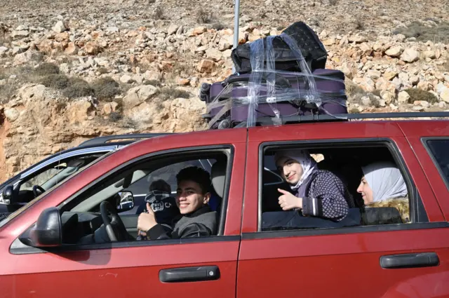 Three young people look at the camera sat inside a red car, two of them putting their thumbs up, and a younger child looking out the car window on the opposite side