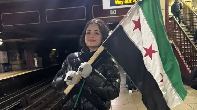 Teenager standing on a London Underground platform wearing a black puffer jacket, grey mittens, and holding the flag of the Syrian opposition