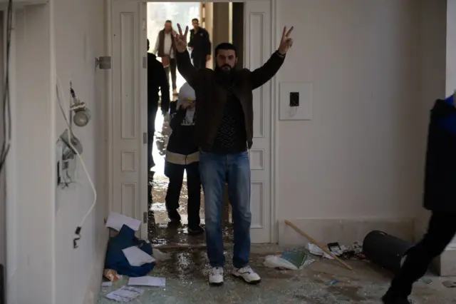 A man holds two fingers up as he walks into a damaged room in the vacated presidential palace