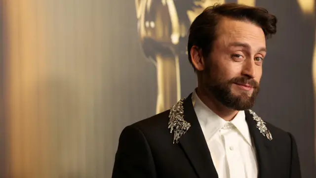 Kieran Culkin smiles at the camera. He wears a black suit with gold decoration on the neck and a white shirt