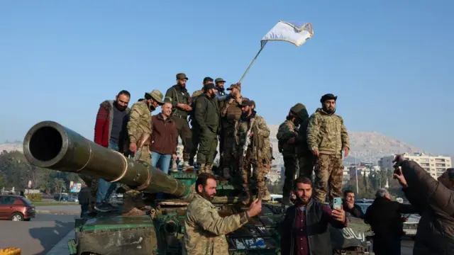 Rebel fighters stand on a tank waving a flag and holding their phones