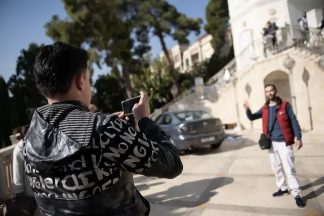 At the front of the picture a man holds a smartphone, taking a picture of another man who poses in front of the presidential palace