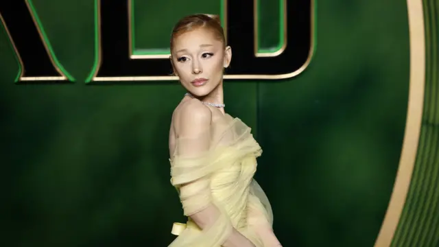 Ariana Grande wears a yellow ball gown and poses for photos in front of a giant green Wicked backdrop