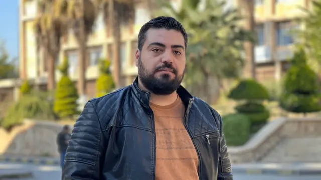 Man looks into camera. He's wearing a black leather jacket and a brown t-shirt. He's standing in front of a tall building with tall palm trees in front of it