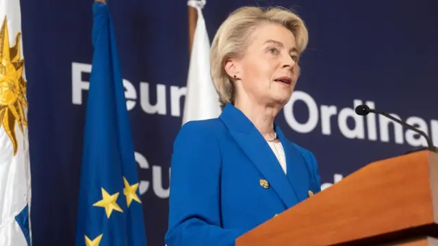 Ursula von der Leyen standing at a podium in a blue suit in front of an EU flag.