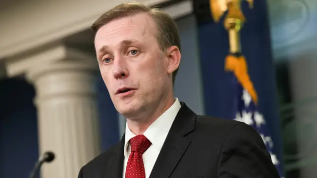 Jake Sullivan wearing a dark suit and red tie stands in front of a United States flag.