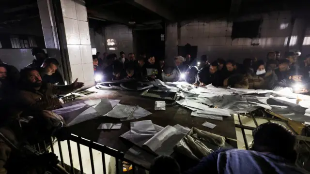 Dozens of people gather around piles of paper with torches inside a room in the prison