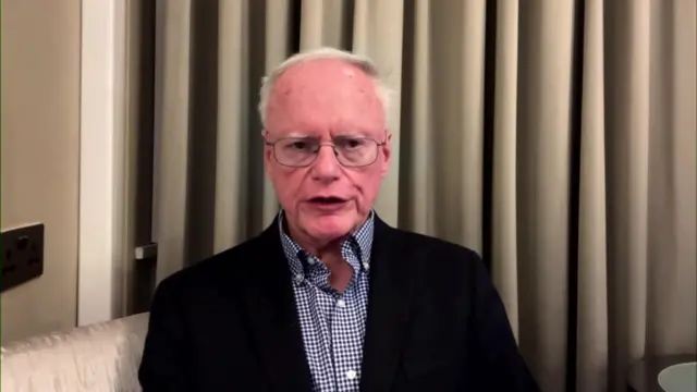 James Jeffrey wearing a chequered shirt with a black blazer and glasses, speaking to the camera