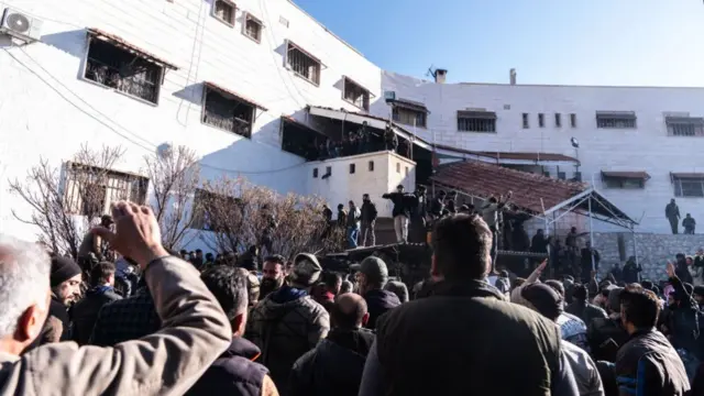 A crowd of people, including men stood on rooftops, outside the prison