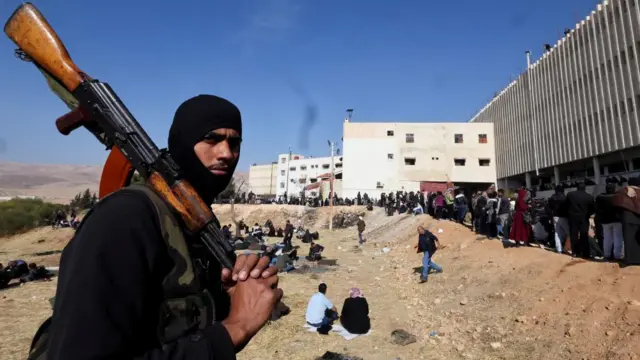 A man holds a gun over his shoulder in front of a queue of people outside the prison