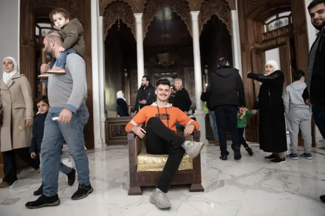 A man in an orange hoodie poses in a chair in the presidential palace while others explore the grounds