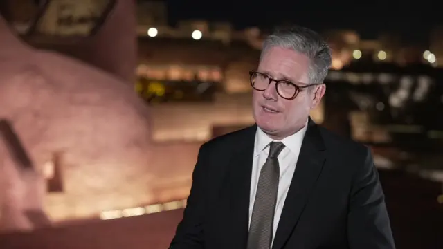 Keir Starmer  wearing a black suit, grey tie and black rimmed glasses talking to the camera